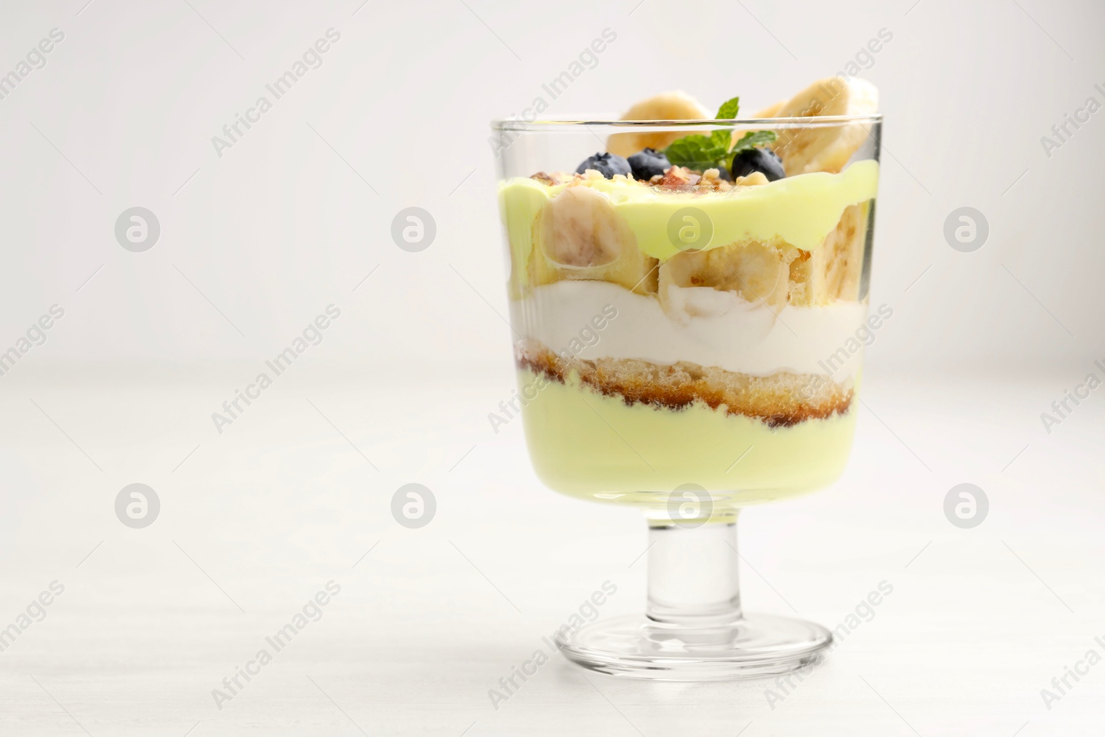 Photo of Tasty trifle dessert. Banana, blueberries, sponge cake and whipped cream in glass on white wooden table, space for text