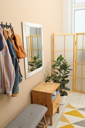 Photo of Coat rack with clothes, mirror, houseplant, shoe storage bench and folding screen in hallway. Interior design