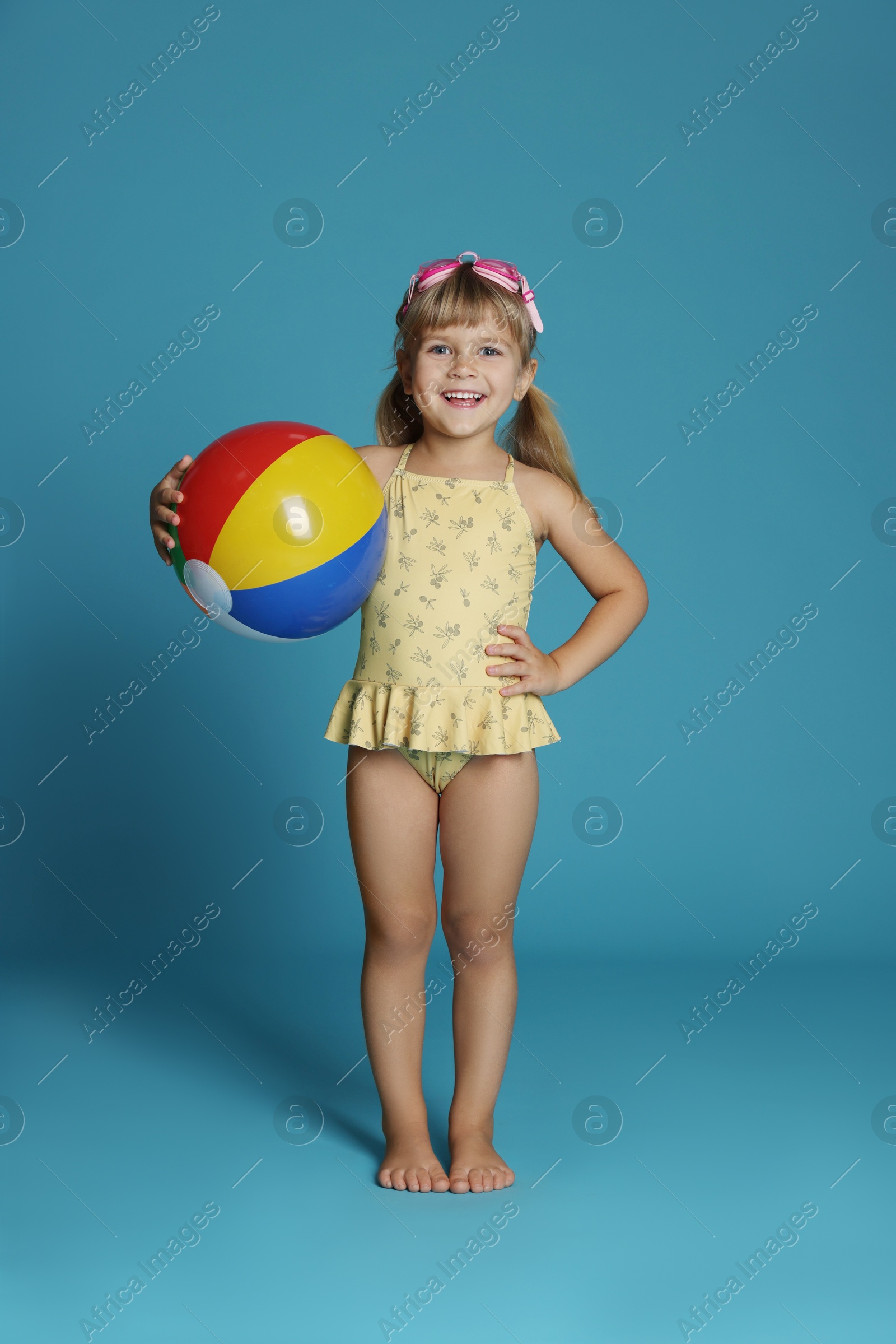 Photo of Cute little girl in swimsuit with inflatable ball and swim goggles on light blue background