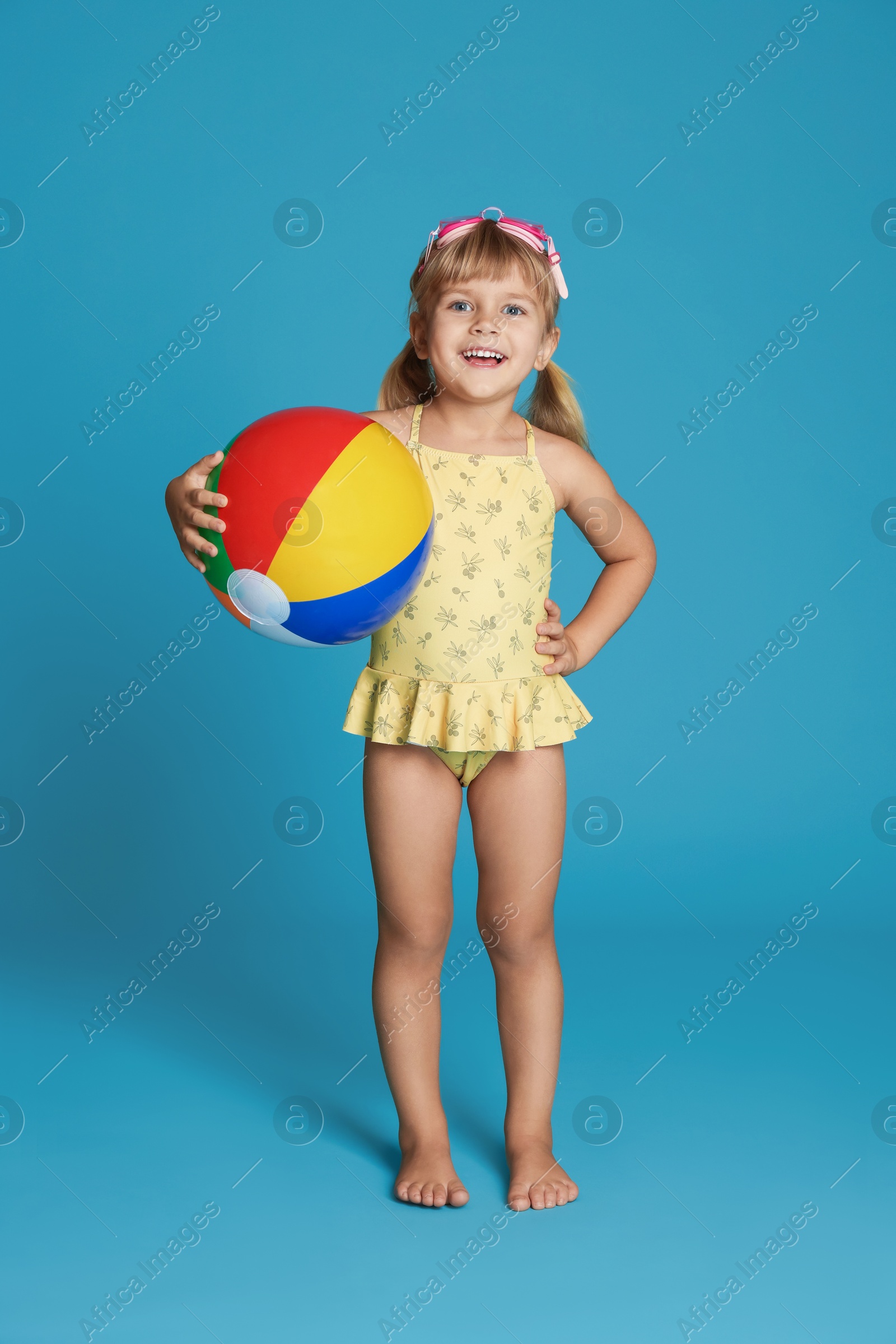 Photo of Cute little girl in swimsuit with inflatable ball and swim goggles on light blue background