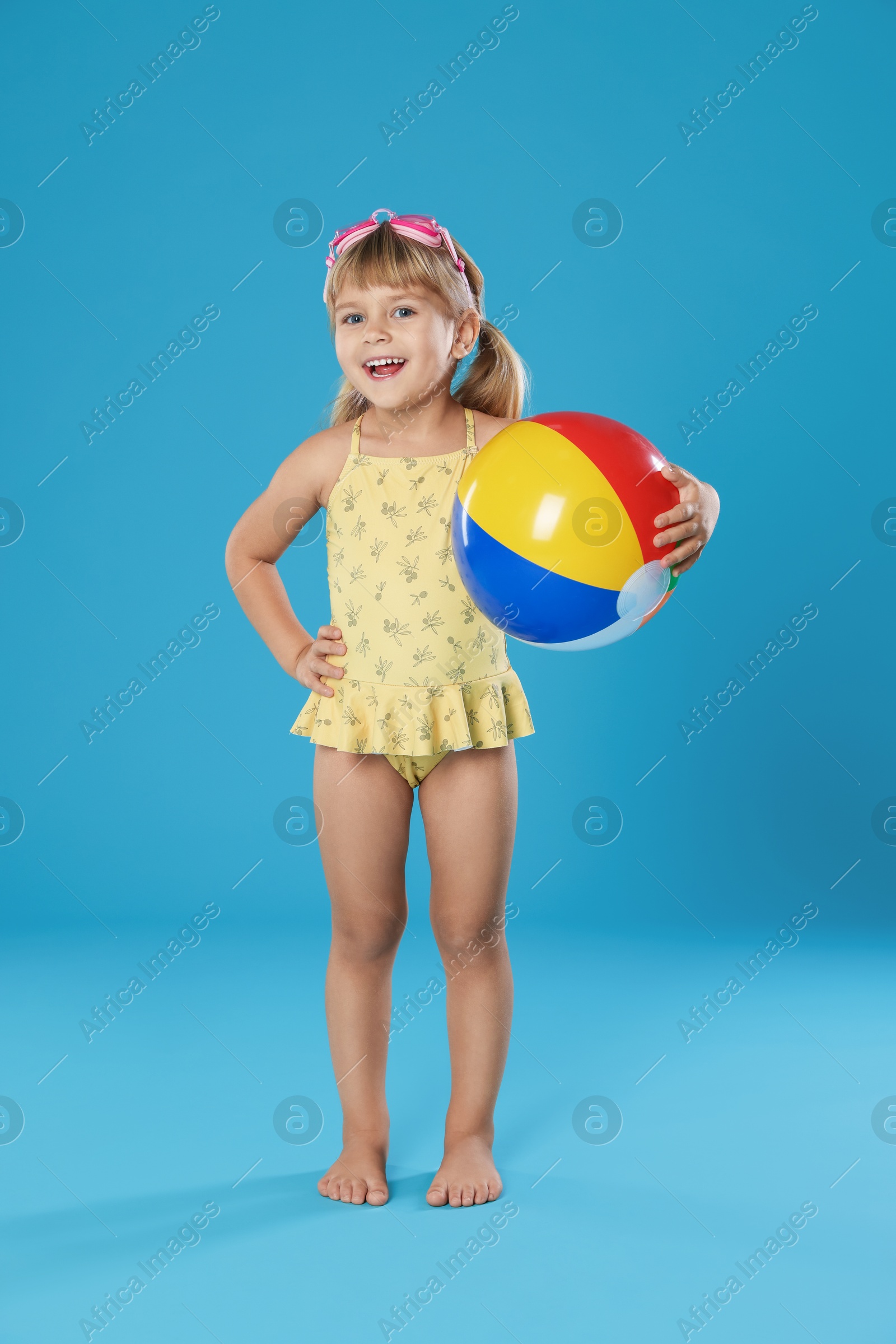 Photo of Cute little girl in swimsuit with inflatable ball and swim goggles on light blue background