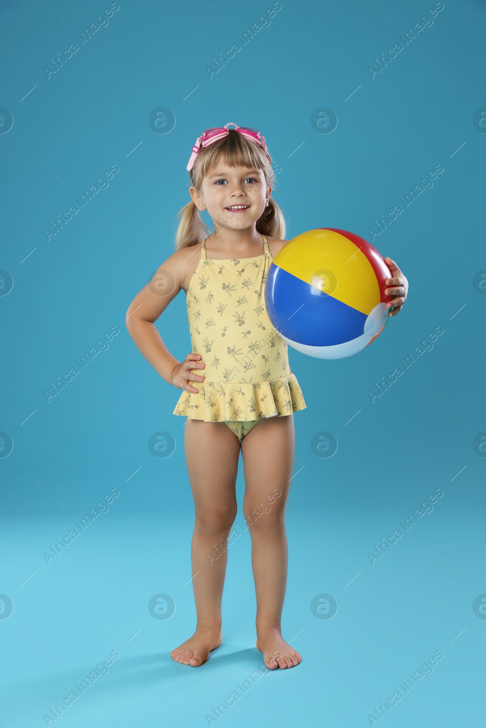 Photo of Cute little girl in swimsuit with inflatable ball and swim goggles on light blue background