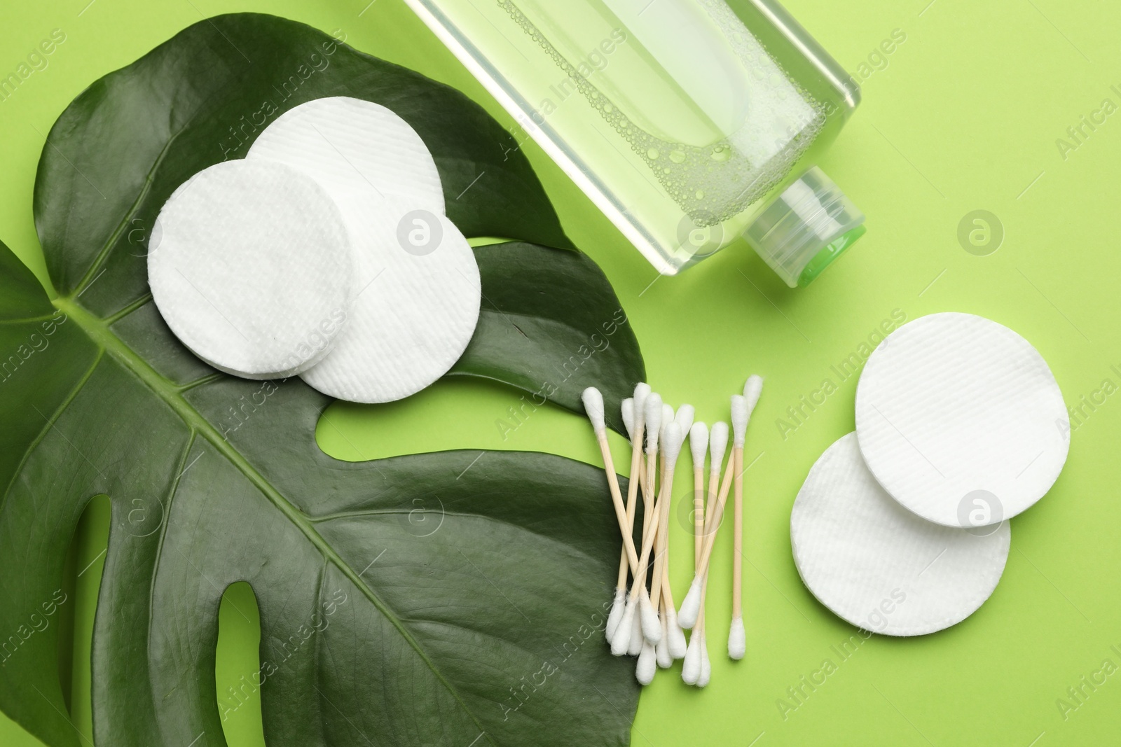 Photo of Cotton pads, swabs, cosmetic product and monstera leaf on green background, flat lay