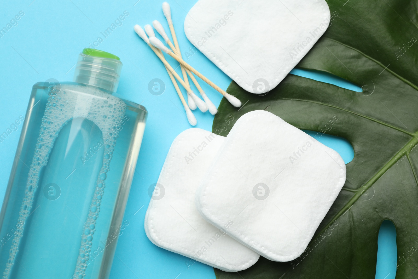 Photo of Cotton pads, swabs, cosmetic product and monstera leaf on light blue background, flat lay