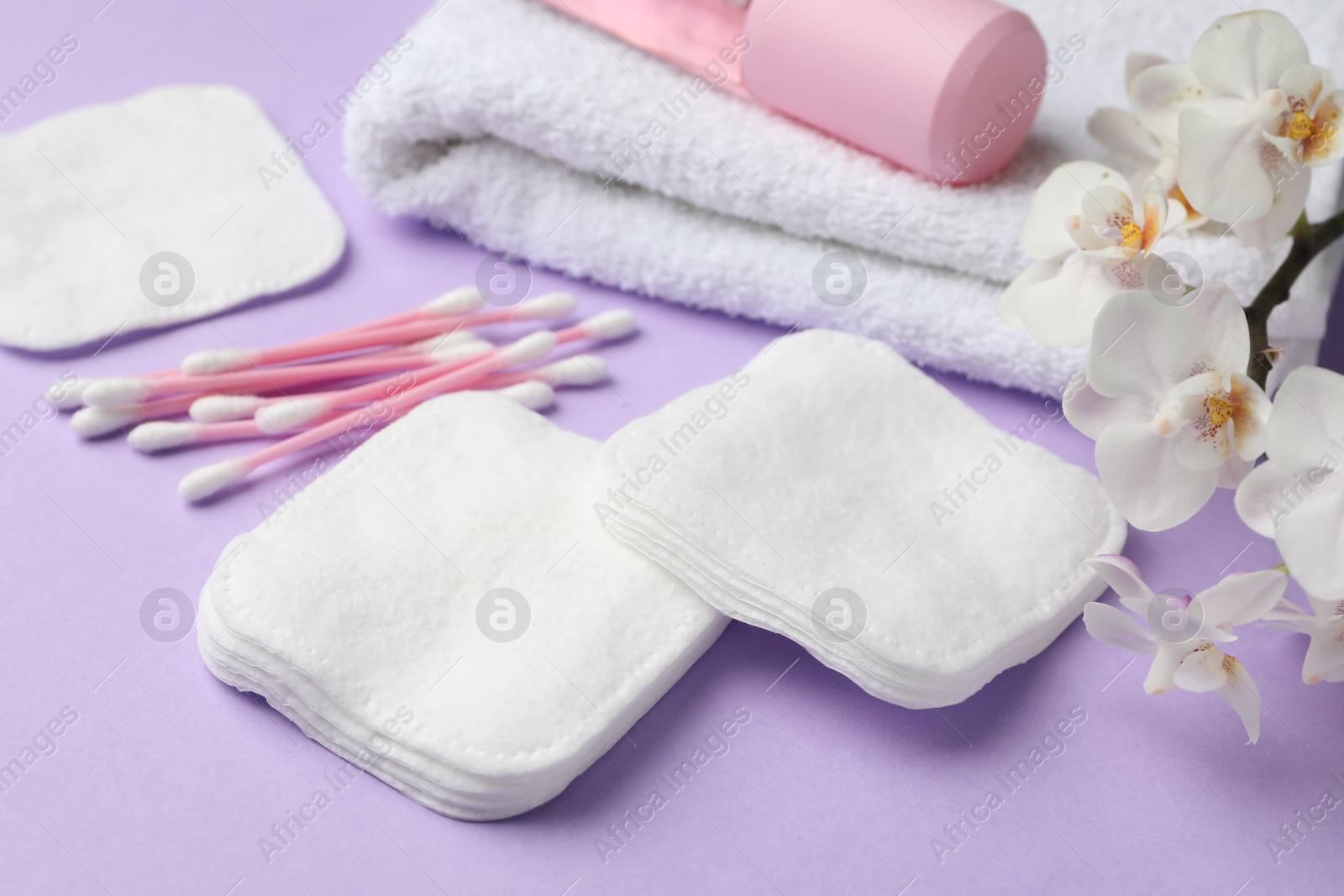 Photo of Cotton pads, swabs, cosmetic product, towel and orchid branch on violet background, closeup