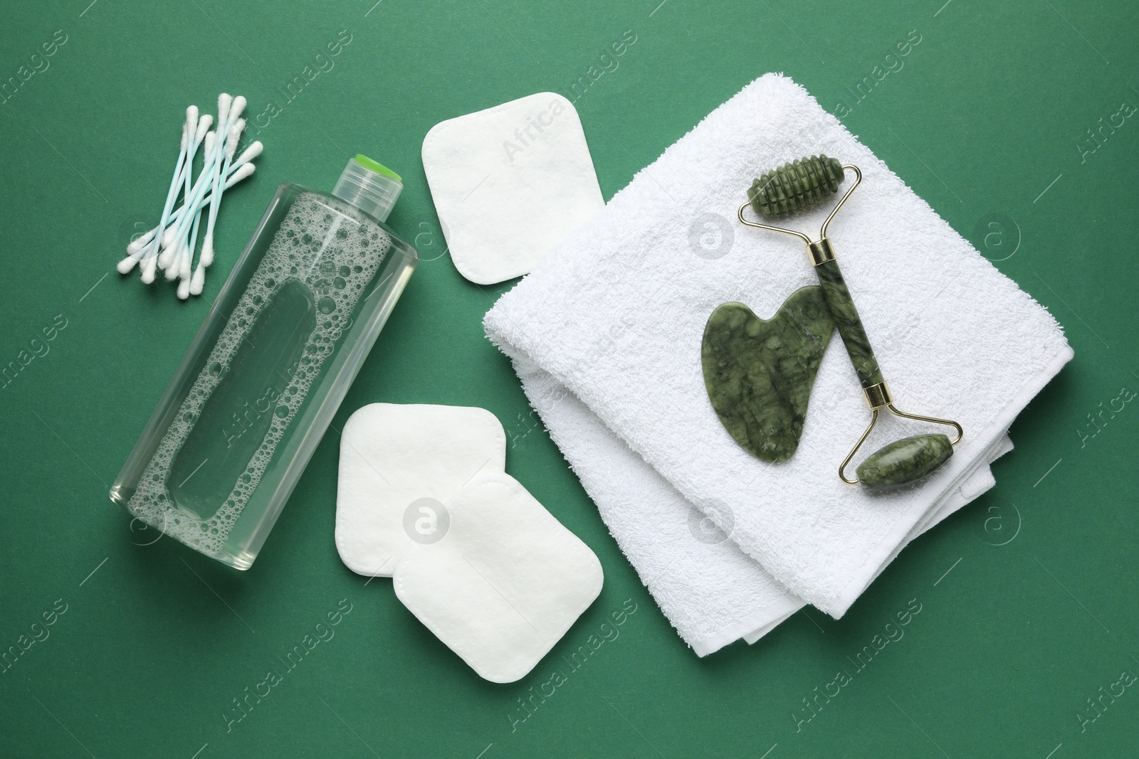 Photo of Cotton pads, swabs, cosmetic product, face roller, gua sha tool and towel on green background, flat lay