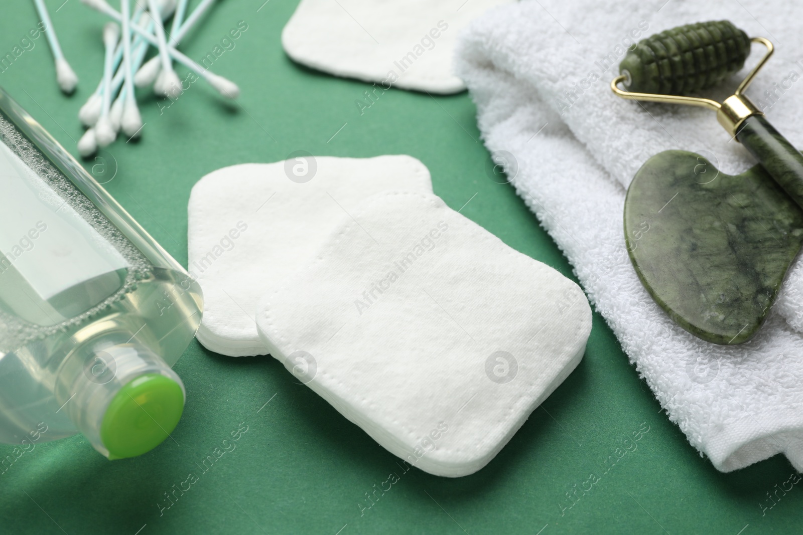 Photo of Cotton pads, swabs, cosmetic product, face roller, gua sha tool and towel on green background, closeup