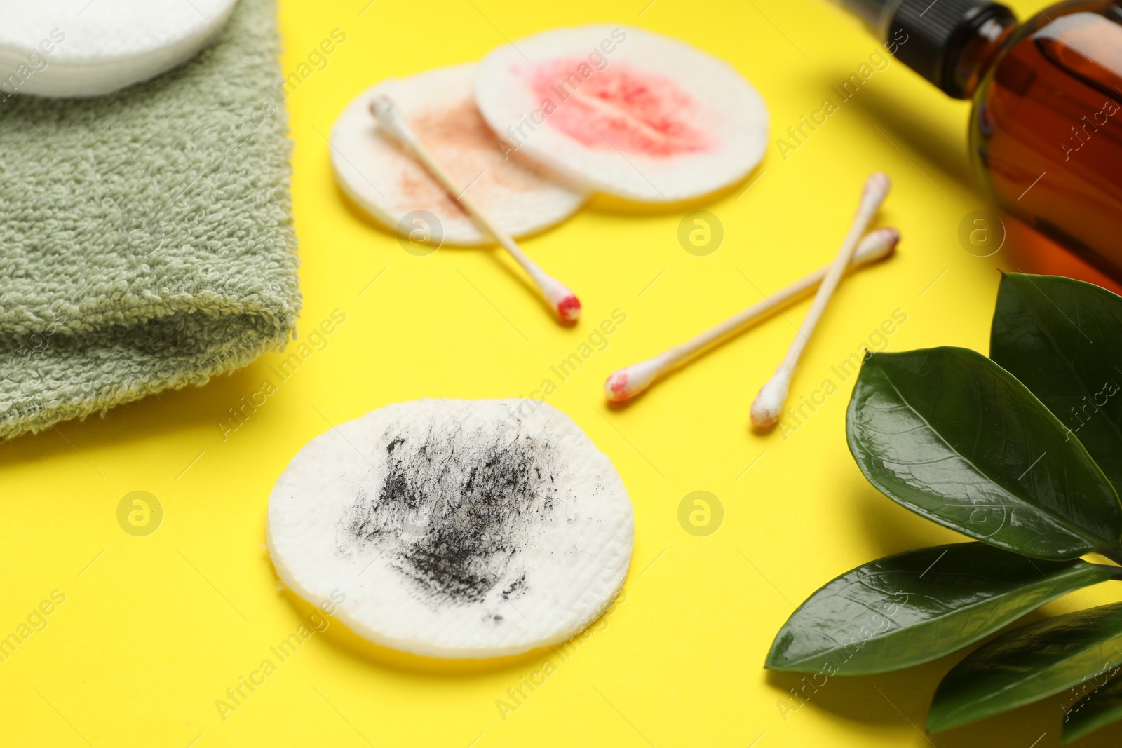 Photo of Dirty cotton pads, swabs, cosmetic product, towel and green leaves on yellow background, closeup