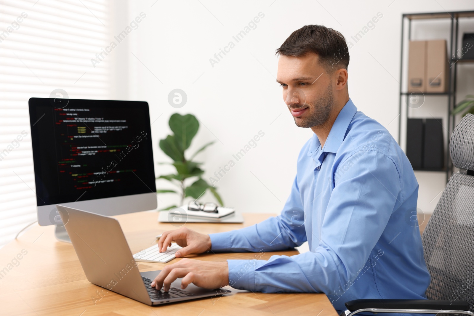 Photo of Programmer working on laptop and computer at wooden desk indoors