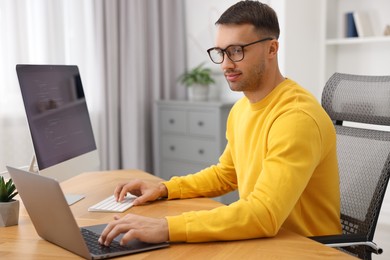 Photo of Programmer working on laptop and computer at wooden desk indoors