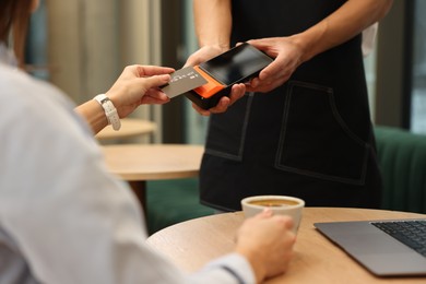 Photo of Woman paying with credit card via terminal at wooden table in cafe, closeup