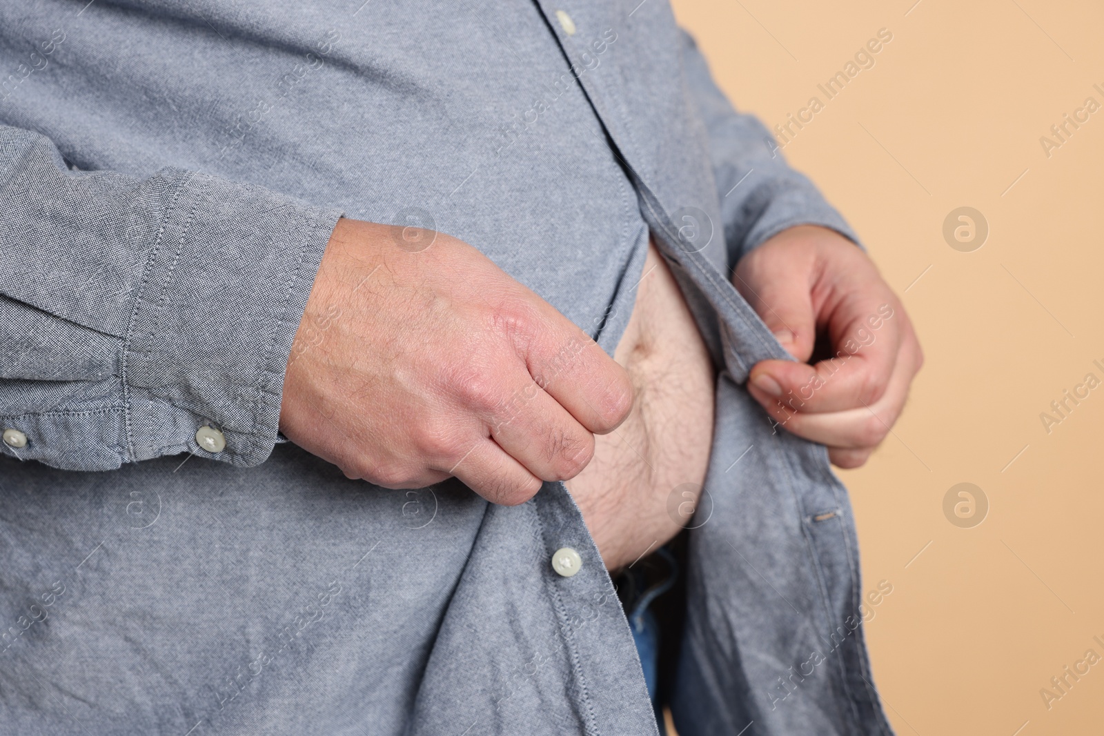 Photo of Overweight man trying to button up tight shirt on beige background, closeup