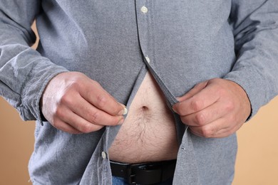 Photo of Overweight man trying to button up tight shirt on beige background, closeup