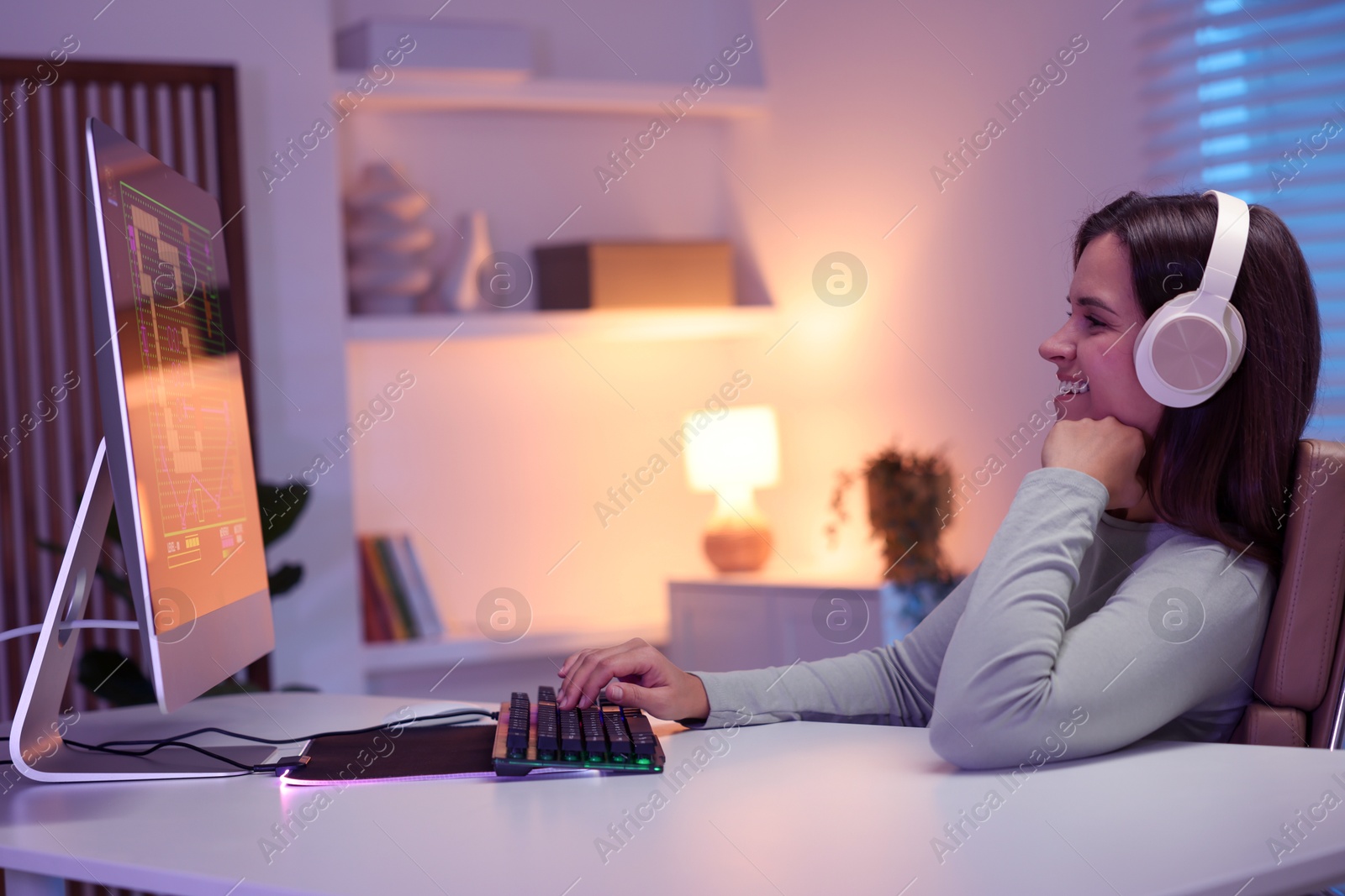 Photo of Smiling woman in headphones playing video game with keyboard at table indoors