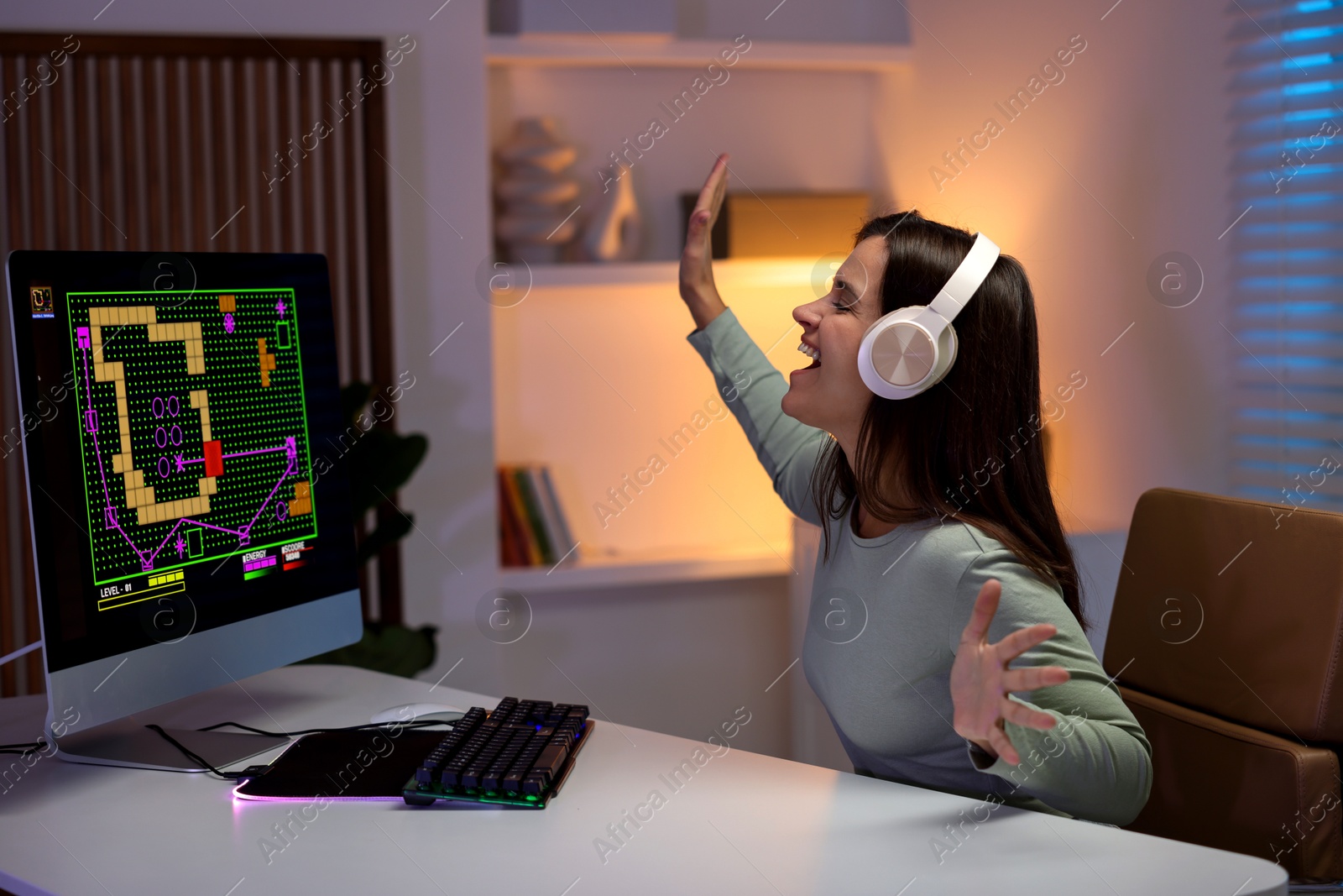 Photo of Happy woman in headphones playing video game at table with keyboard and monitor indoors