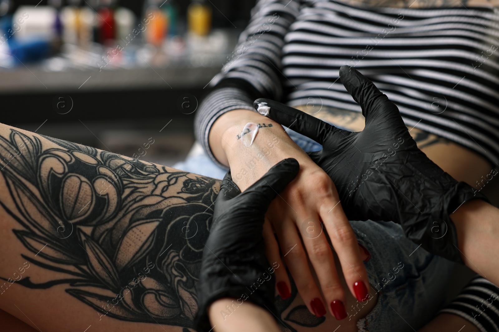 Photo of Professional artist in gloves applying cream onto woman's hand with fresh tattoo in salon, closeup