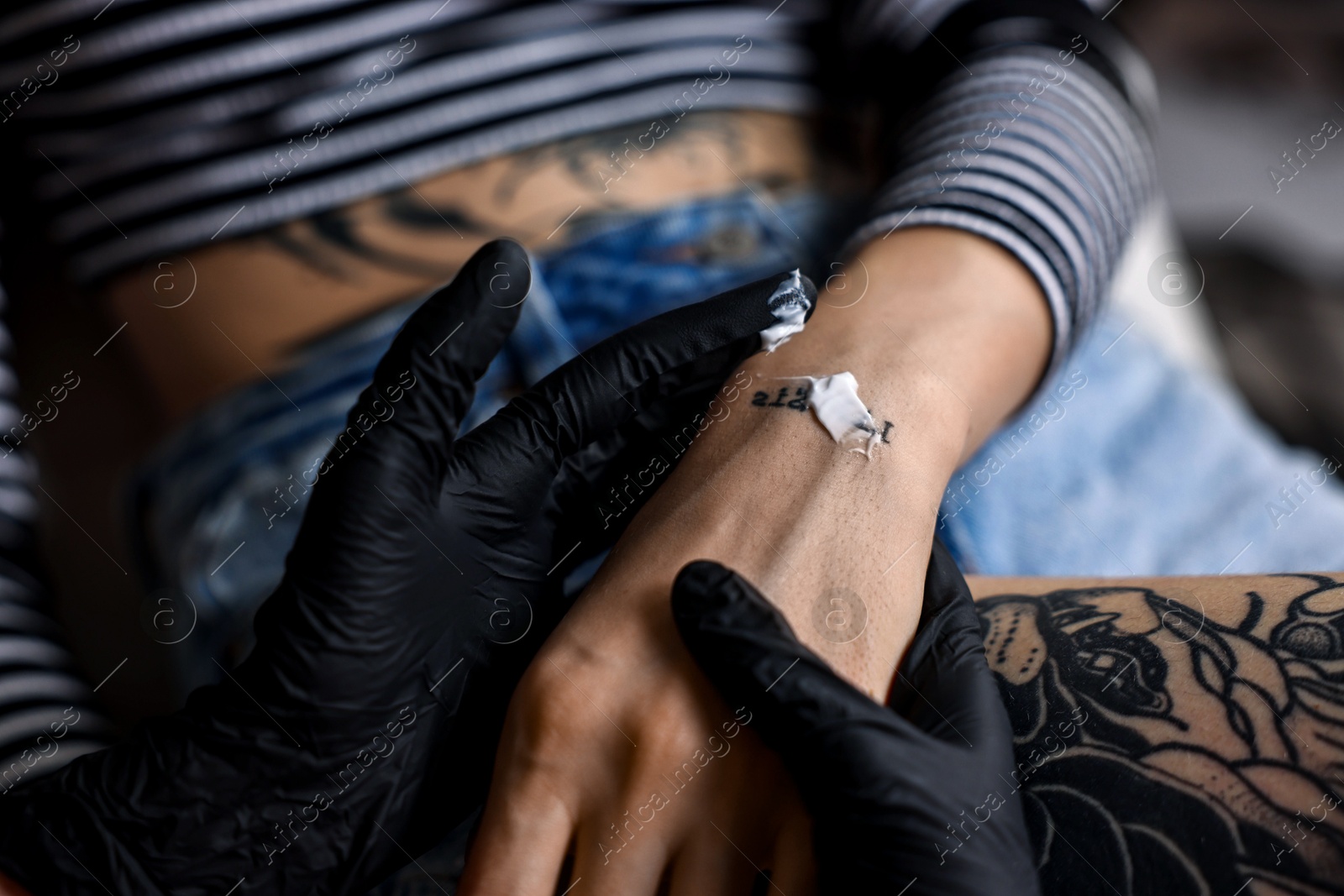 Photo of Professional artist in gloves applying cream onto woman's hand with fresh tattoo in salon, closeup