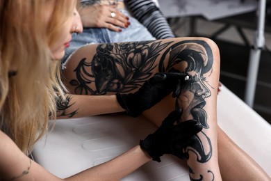 Photo of Professional artist in gloves applying cream onto woman's leg with fresh tattoo in salon, closeup