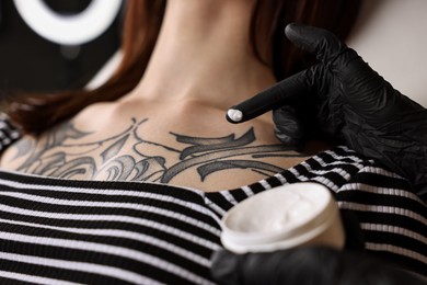 Photo of Professional artist in gloves applying cream onto woman's skin with fresh tattoo in salon, closeup