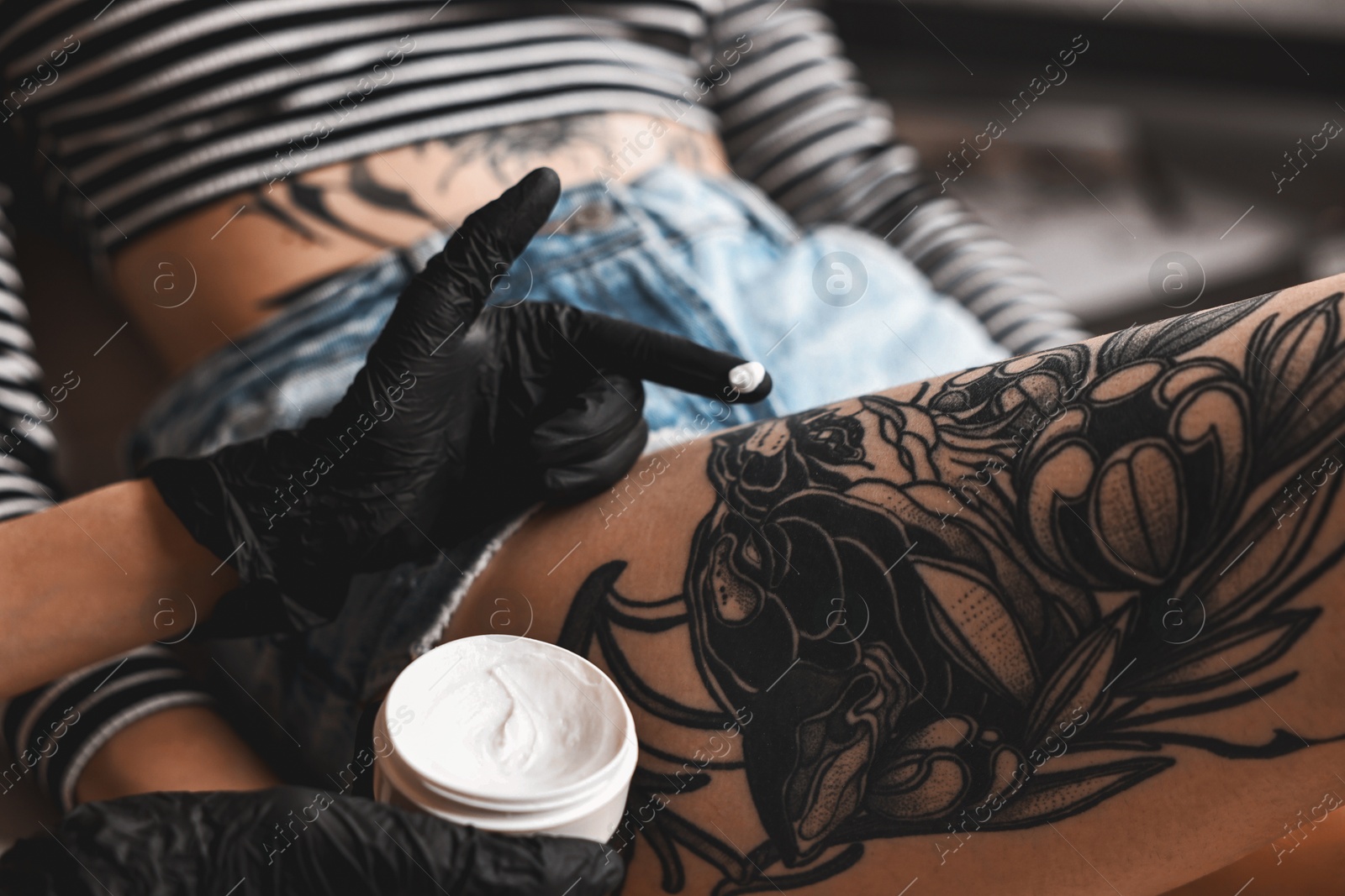 Photo of Professional artist in gloves applying cream onto woman's leg with fresh tattoo in salon, closeup