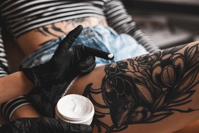 Photo of Professional artist in gloves applying cream onto woman's leg with fresh tattoo in salon, closeup