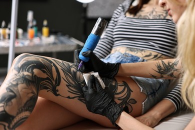 Photo of Professional artist tattooing woman's leg with machine in salon, closeup