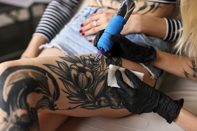 Photo of Professional artist tattooing woman's leg with machine in salon, closeup