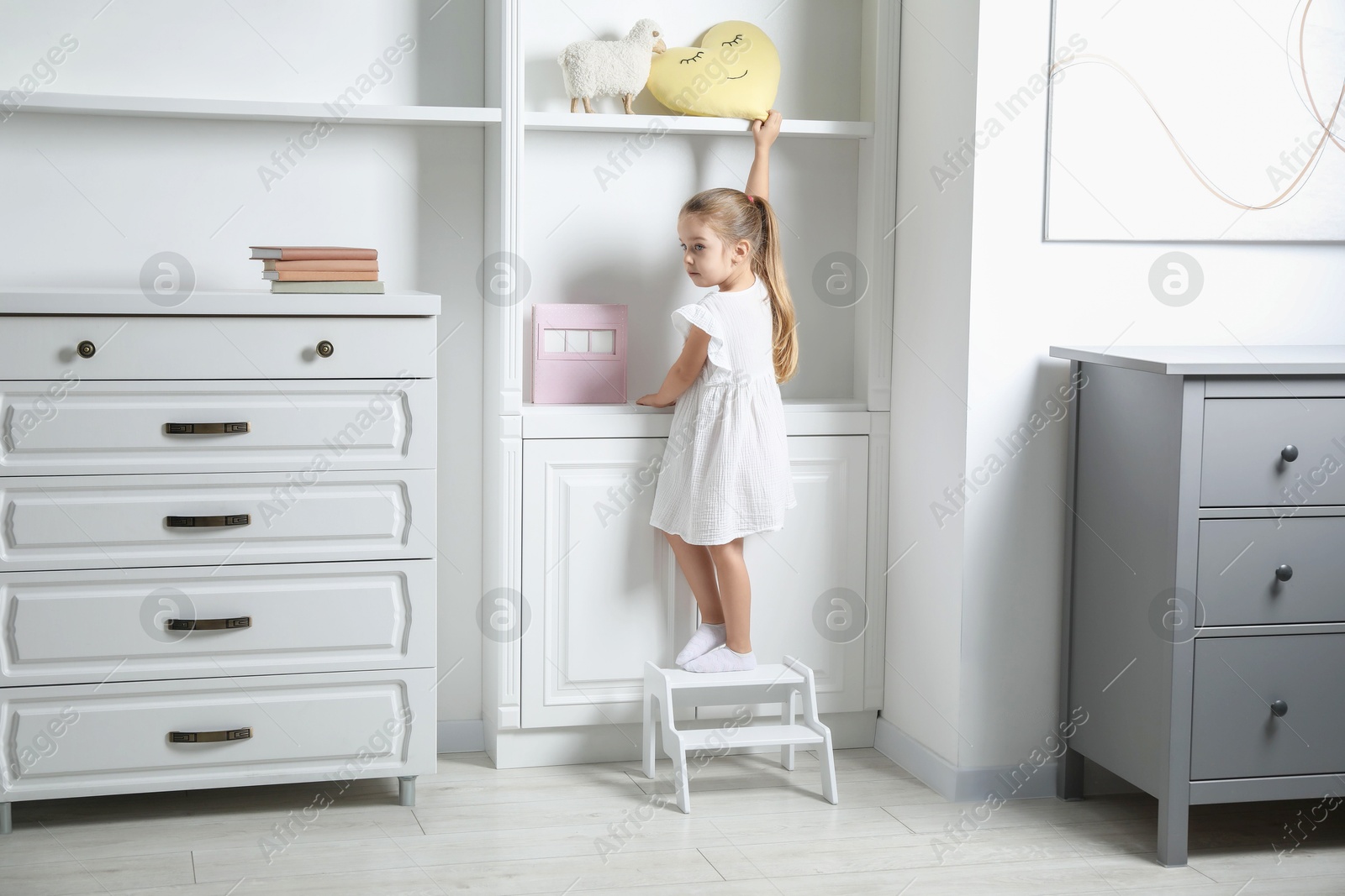 Photo of Little girl standing on step stool and reaching for toys on shelf indoors