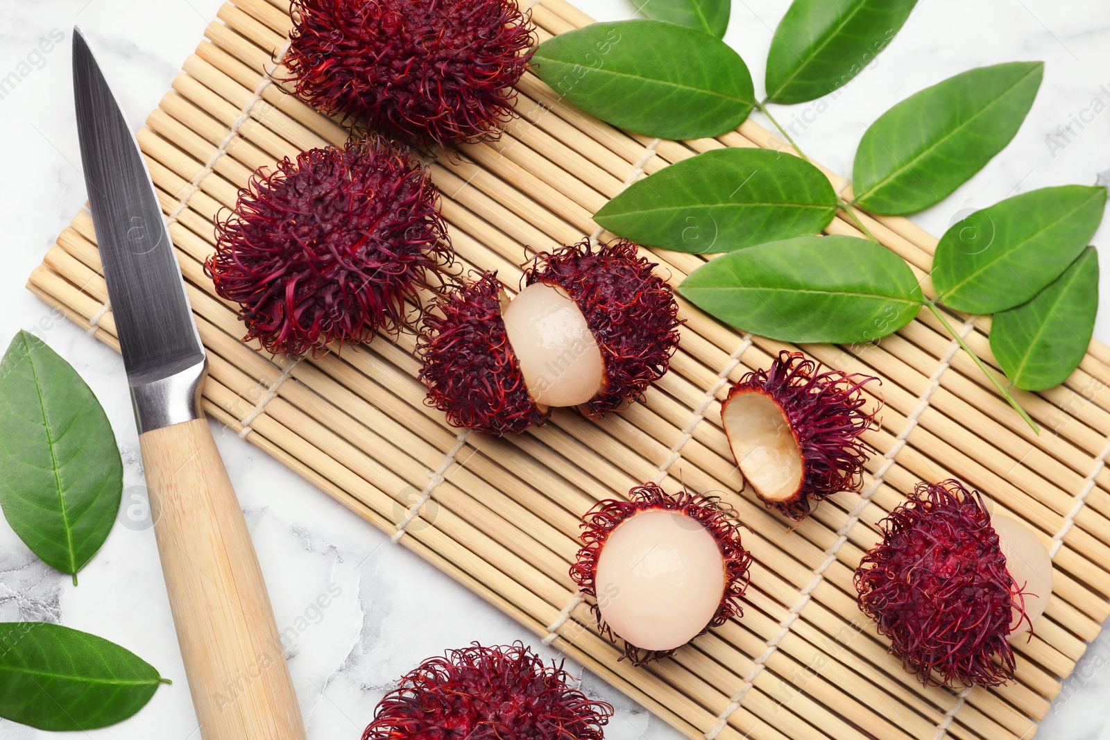 Photo of Delicious ripe rambutans, knife and green leaves on white marble table, flat lay