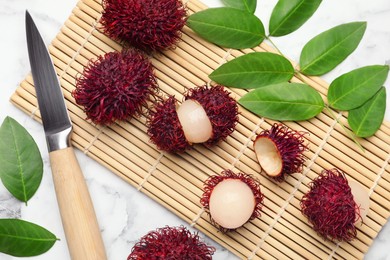 Photo of Delicious ripe rambutans, knife and green leaves on white marble table, flat lay