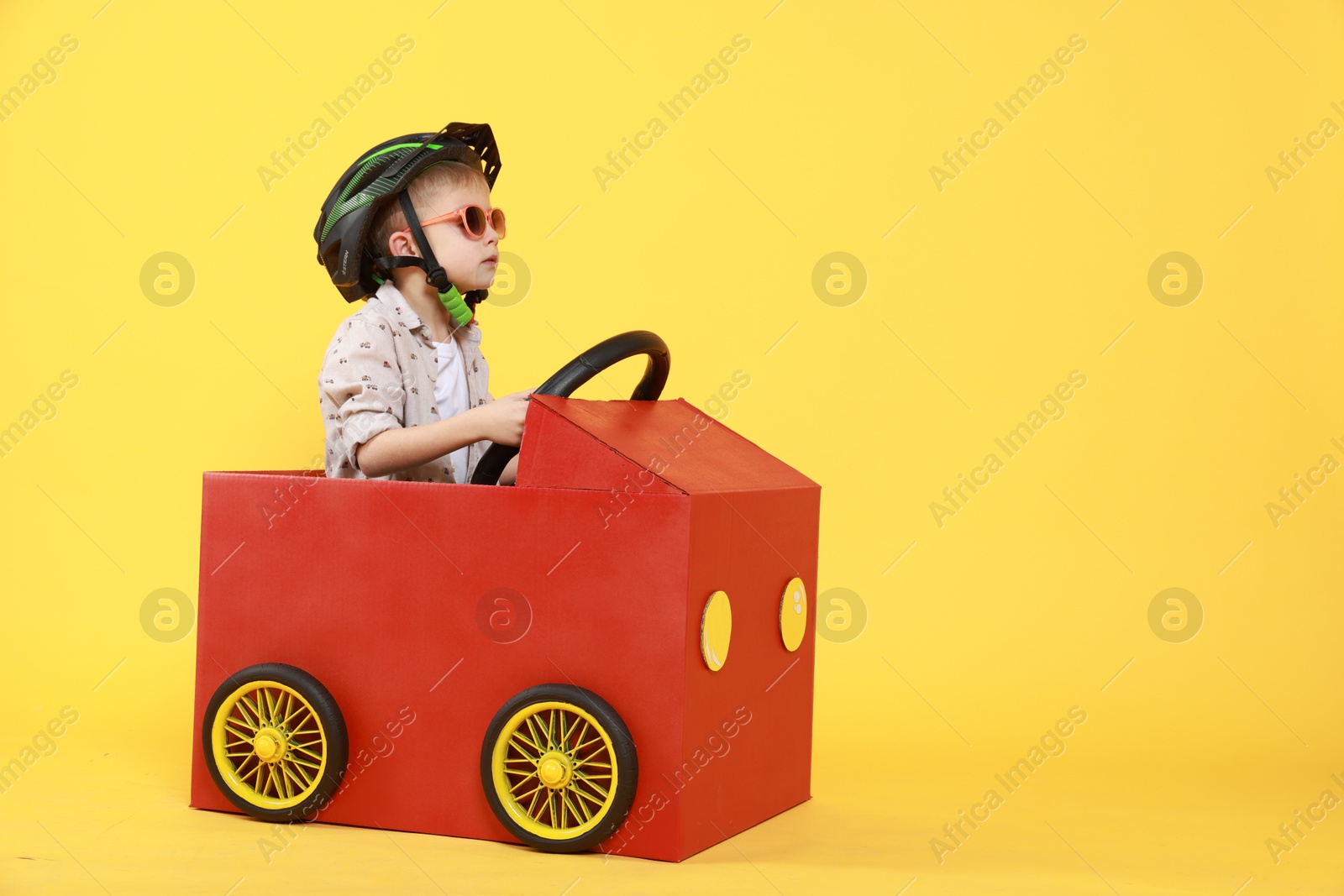 Photo of Little boy driving car made of cardboard on yellow background. Space for text