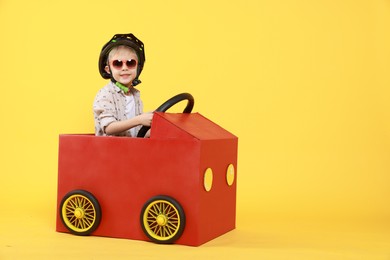 Photo of Little boy driving car made of cardboard on yellow background. Space for text