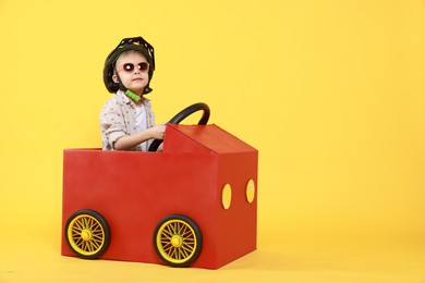 Photo of Little boy driving car made of cardboard on yellow background. Space for text