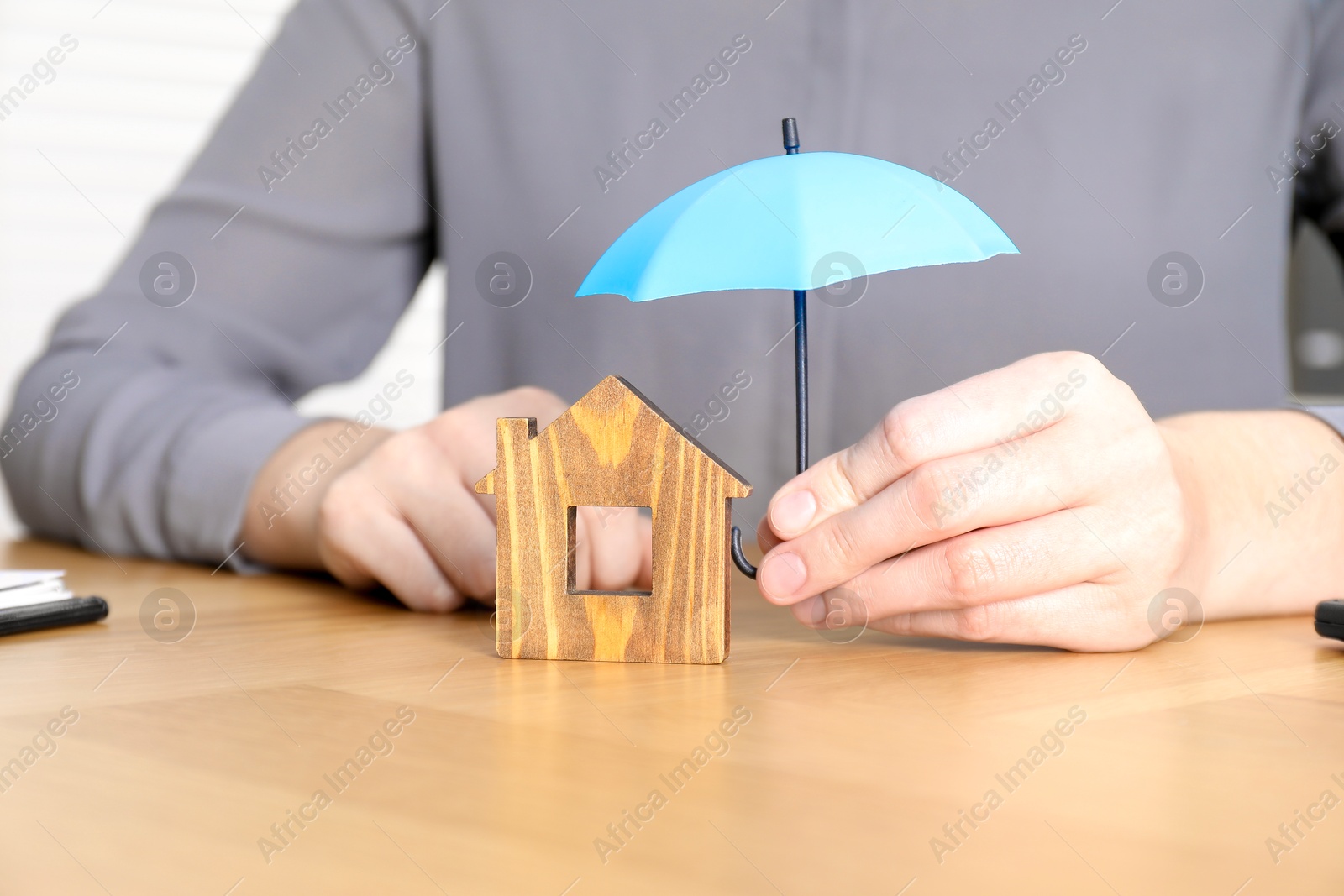 Photo of Property insurance. Real estate agent holding small umbrella above wooden house figure at table indoors, closeup