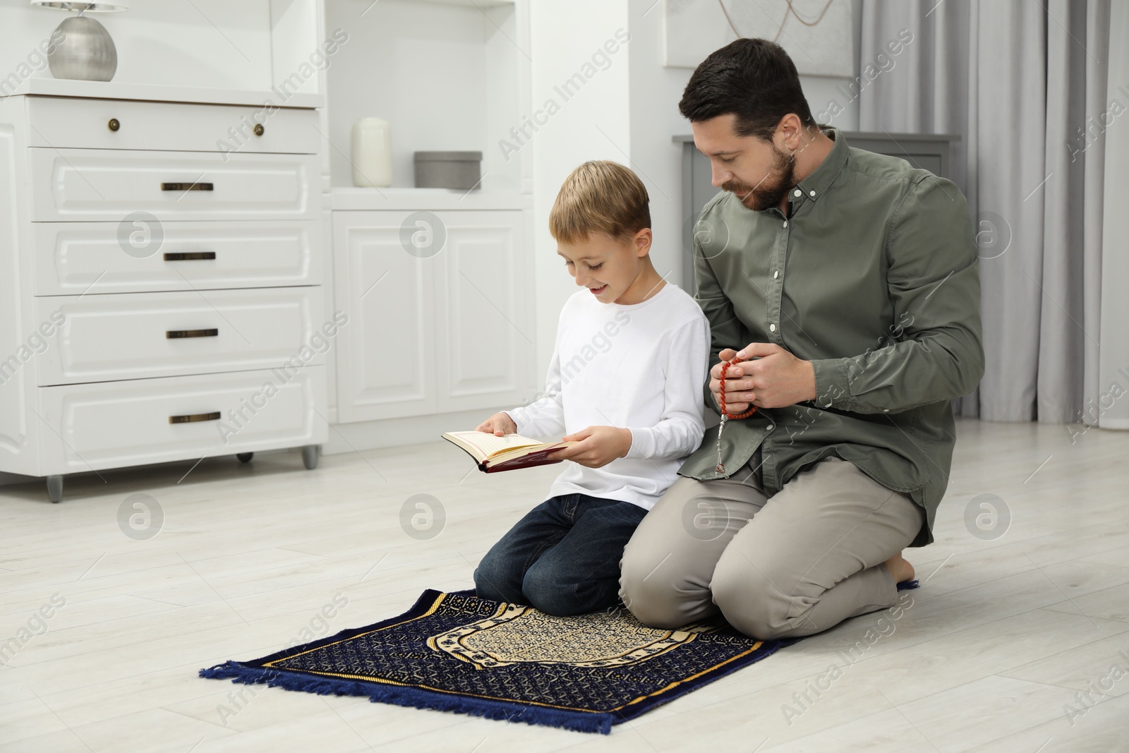 Photo of Muslim man with his son reading Quran and praying on mat at home