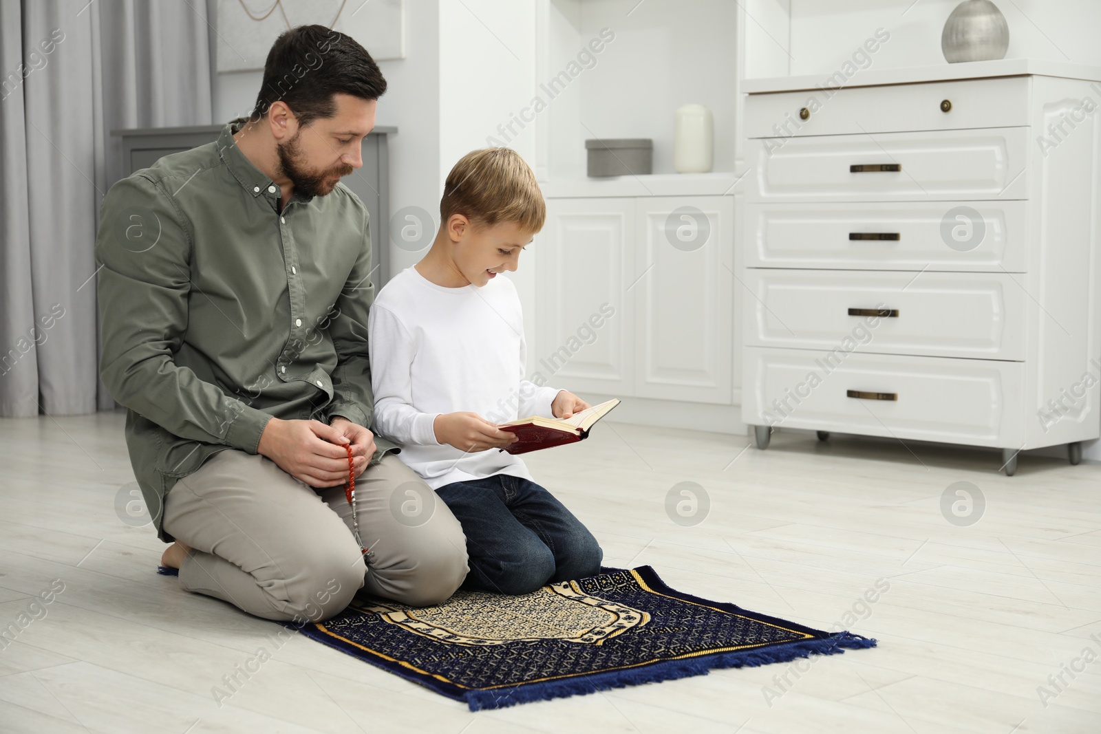 Photo of Muslim man with his son reading Quran and praying on mat at home, space for text