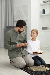 Photo of Muslim man with his son reading Quran and praying on mat at home