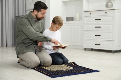 Photo of Muslim man with his son reading Quran and praying on mat at home, space for text