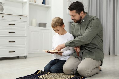 Photo of Muslim man with his son reading Quran and praying on mat at home, space for text