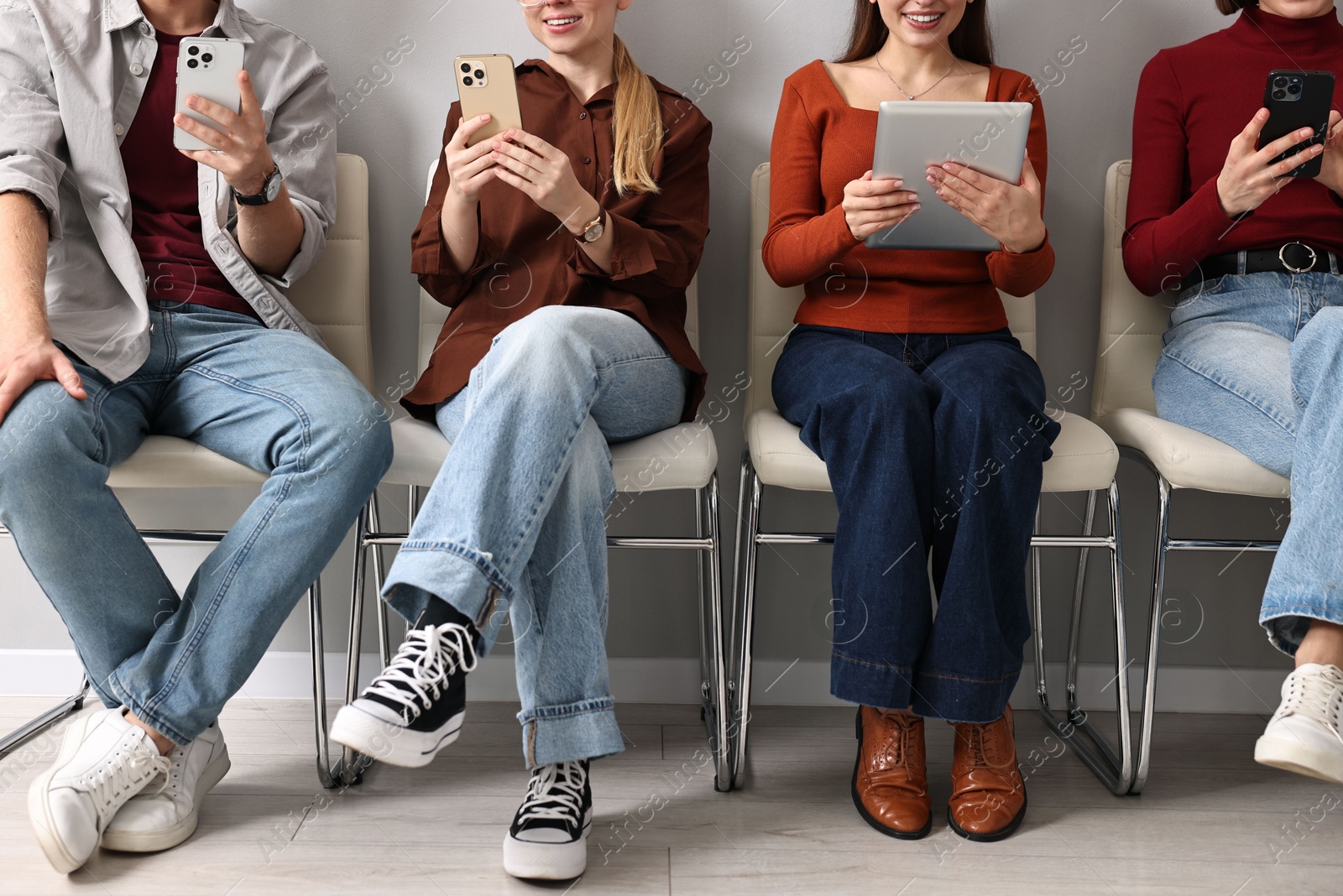 Photo of Group of people using different gadgets on chairs near grey wall indoors, closeup. Modern technology