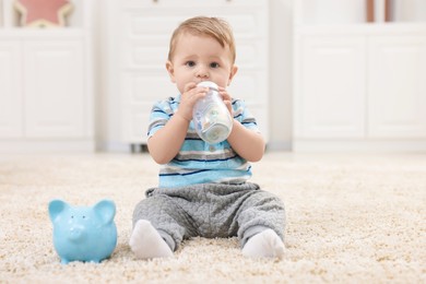 Photo of Cute little baby with money in bottle and piggybank on floor at home