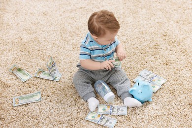 Photo of Little baby with bottle, money and piggybank on floor at home