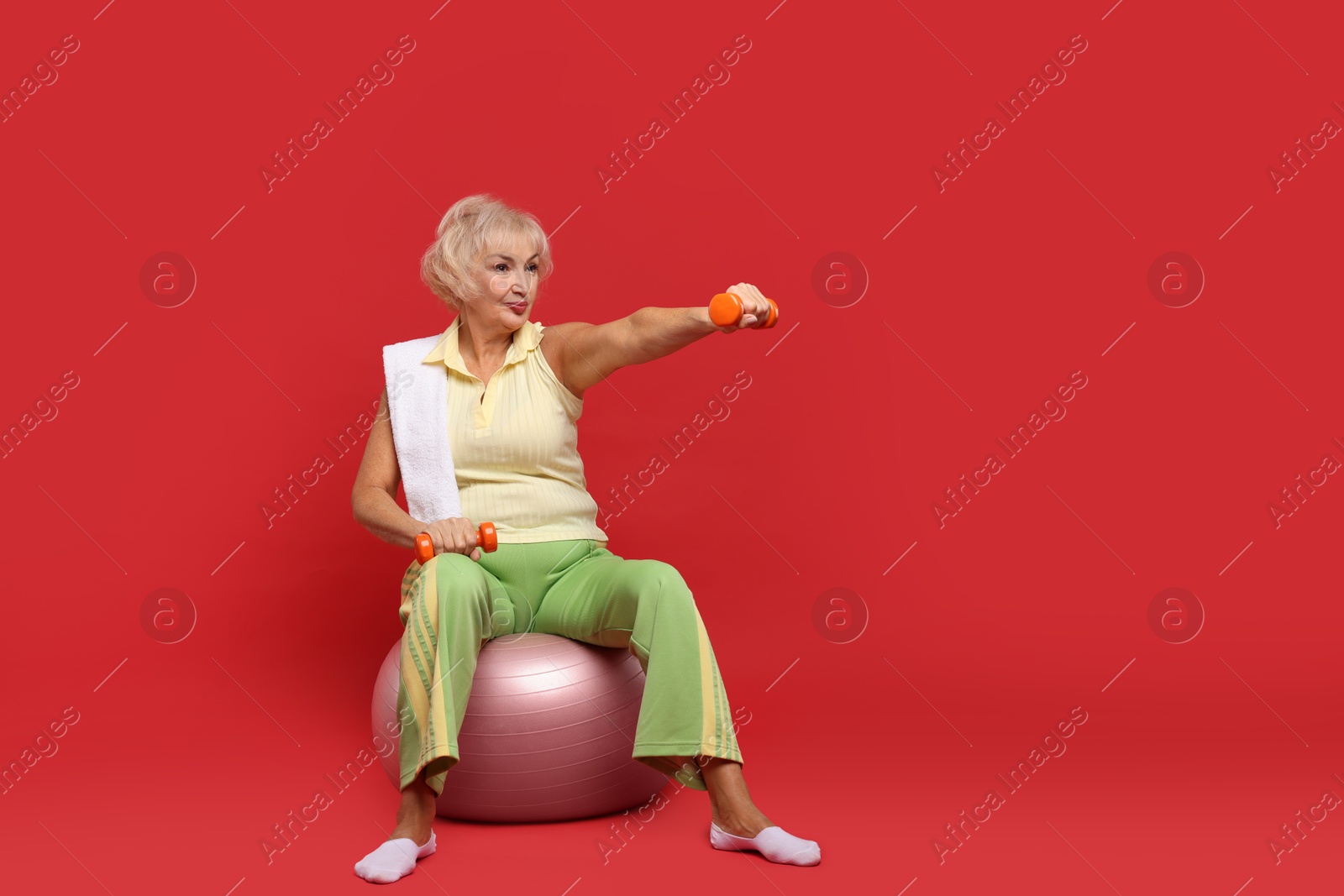 Photo of Senior woman exercising with fitness ball, towel and dumbbells on red background, space for text