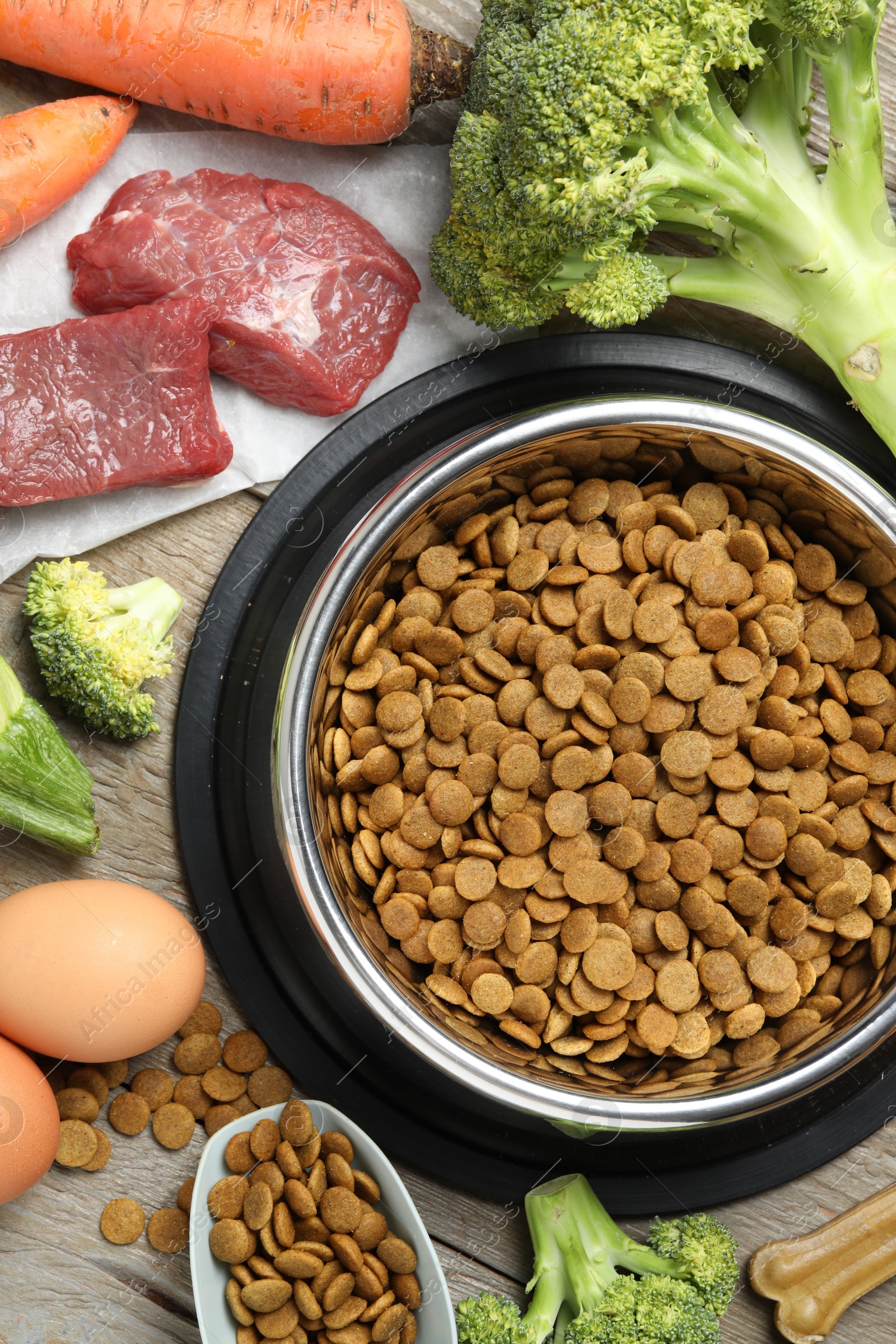 Photo of Dry pet food in feeding bowl among natural products on wooden table, flat lay