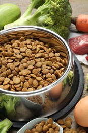 Photo of Dry pet food in feeding bowl among natural products on wooden table, closeup