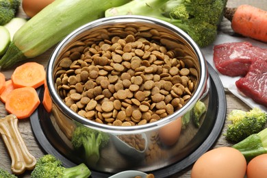 Photo of Dry pet food in feeding bowl among natural products on wooden table, closeup