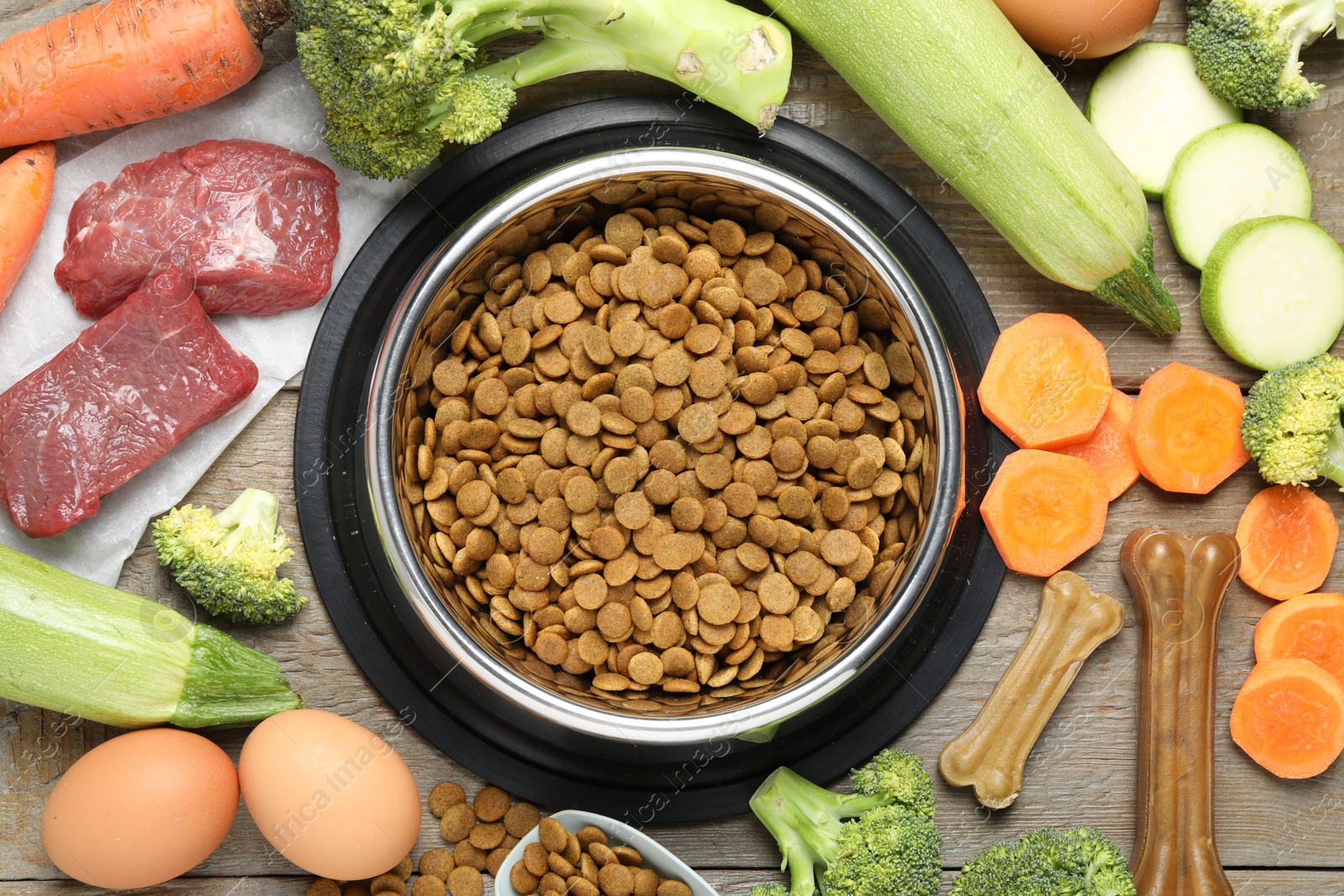 Photo of Dry pet food in feeding bowl among natural products and chew bones on wooden table, flat lay