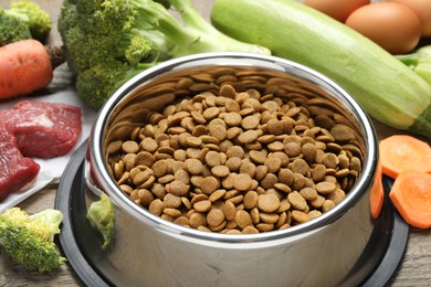 Photo of Dry pet food in feeding bowl among natural products on wooden table, closeup