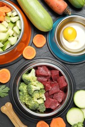 Photo of Natural pet food in feeding bowls and products on dark textured table, flat lay