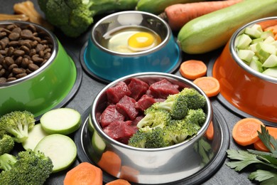 Photo of Natural and dry pet food in feeding bowls among products on dark textured table, closeup